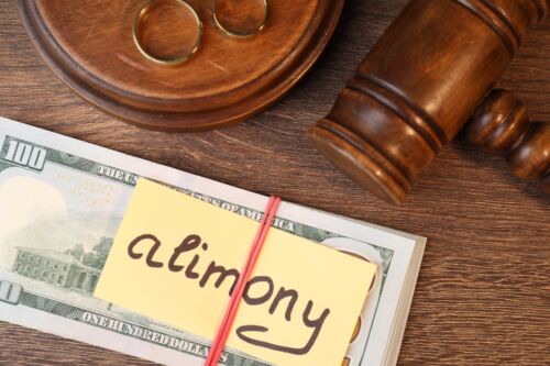 Card with word Alimony, dollar banknotes, gavel and rings on wooden table Visual concept for blog discussing alimony modification.