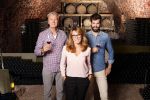 Portrait of multigenerational winery owner family standing at wine cellar. Senior winemaker and young sommelier standing at background and holding in hands a glass of red wine while middle age businesswoman looking at camera and smiling. Concept for inherited family business.