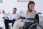 Sad woman sitting on a wheelchair, in the background her husband sitting on a sofa visual concept for legal blog discussing the factors to consider when divorcing a disabled spouse.